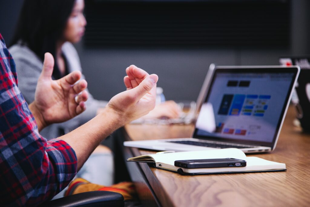View of man's arms and hands, held up in conversation while on a call through a laptop that is front of him on a work table. A woman can be seen in the background also working. Showcases the Senior Technical Leader role listed under CloudAssist jobs and CloudAssist hiring opportunities.